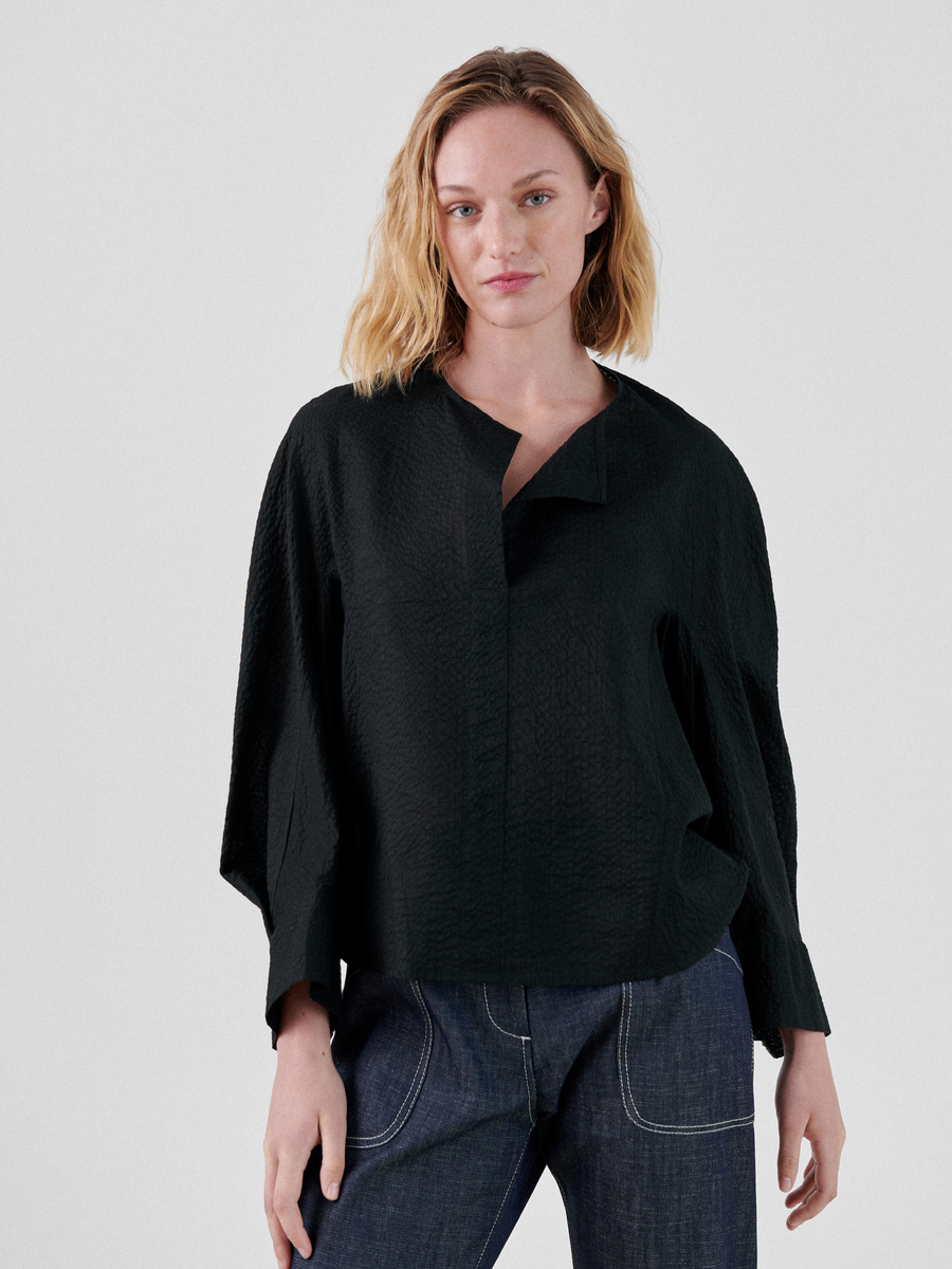 A person with shoulder-length blonde hair is wearing a loose-fitting Long-Sleeved Akeo Shirt by Zero + Maria Cornejo and blue jeans. They are standing against a plain white background, looking directly at the camera with a neutral expression.