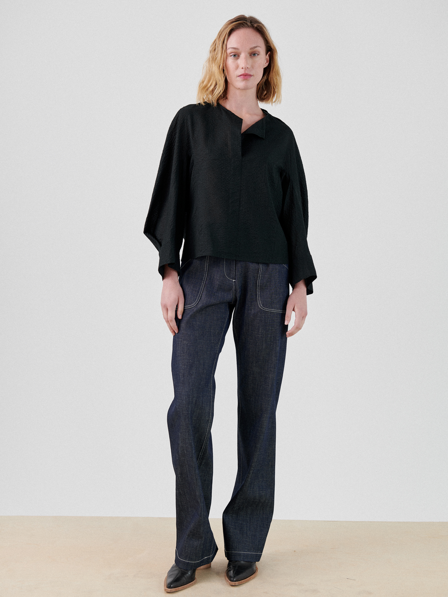 A person with shoulder-length blonde hair is standing against a plain background. They are wearing a loose-fitting black top with wide sleeves and straight-leg dark blue jeans. The Long-Sleeved Akeo Shirt by Zero + Maria Cornejo drapes elegantly on their frame. The person is also wearing black shoes and has a neutral expression.