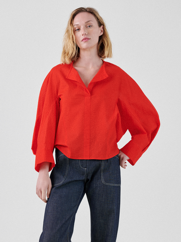 A woman with shoulder-length blonde hair is wearing a bright red Long-Sleeved Akeo Shirt by Zero + Maria Cornejo and black high-waisted jeans. Her stance is relaxed, with one hand resting by her side and the other slightly bent. The background is plain white.