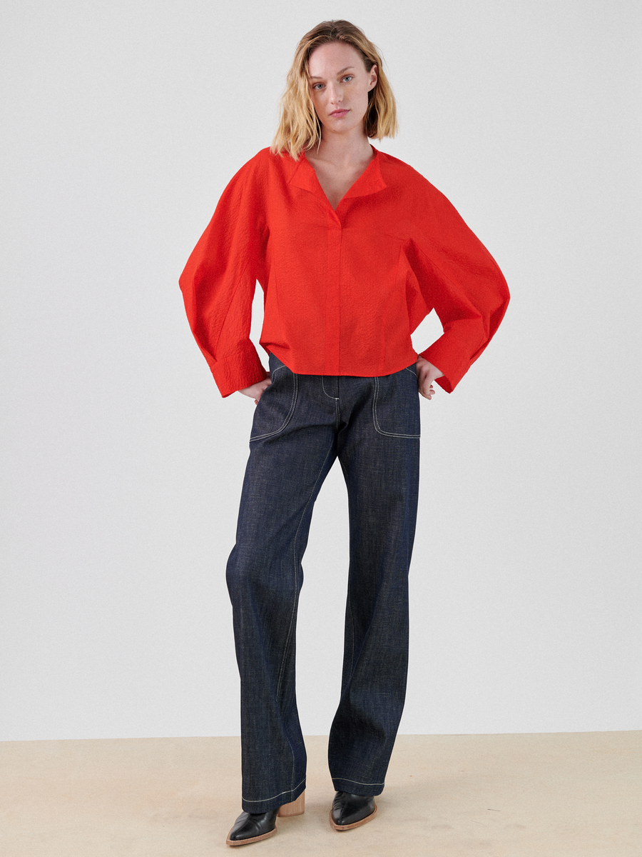 A person with shoulder-length blond hair is standing confidently with hands in pockets. They are wearing a bright red, textured Long-Sleeved Akeo Shirt from Zero + Maria Cornejo, dark wide-leg jeans with white stitching, and black pointed-toe shoes against a plain white background.