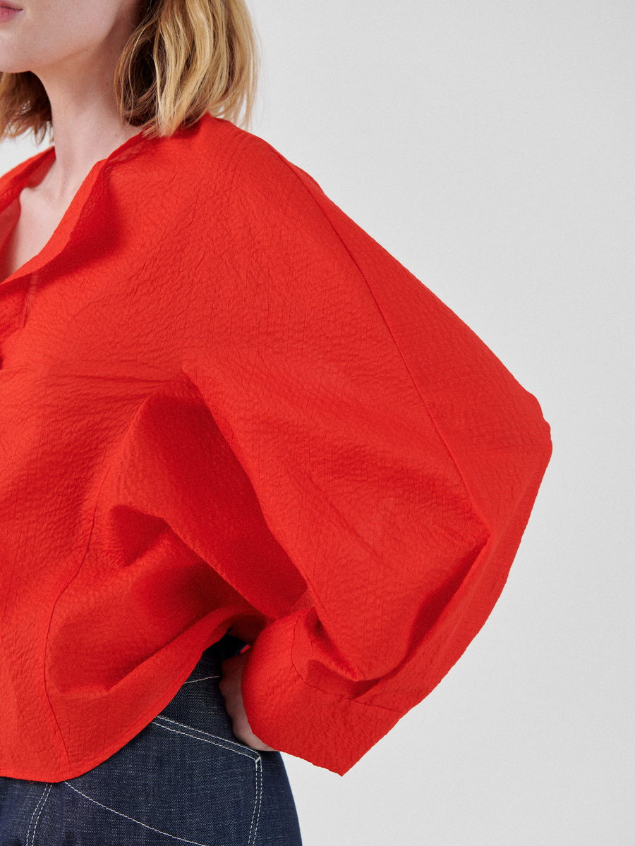 A person in a bright red, textured **Zero + Maria Cornejo Long-Sleeved Akeo Shirt** with oversized, balloon sleeves is partially visible from the shoulders to the mid-waist. They have shoulder-length blonde hair. The background is plain light grey, and the person is wearing dark blue pants.