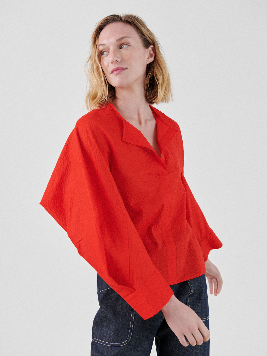 A person with shoulder-length blonde hair is wearing a bright red, Long-Sleeved Akeo Shirt by Zero + Maria Cornejo and dark blue jeans. They are standing and looking to the side against a plain light gray background.