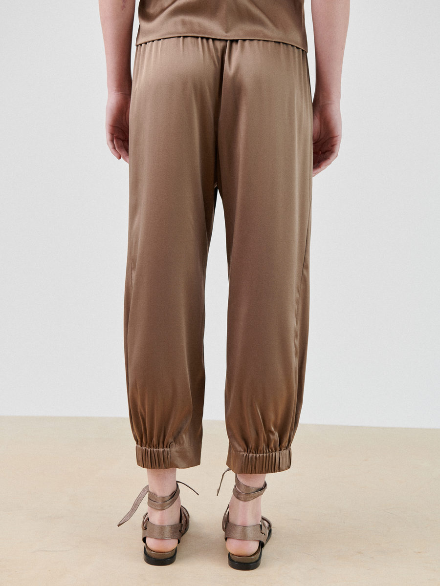 A person wearing Zero + Maria Cornejo Abi Pant in brown satin stretch silk charmeuse with an elastic waistband and elastic cuffs at the ankles is standing. The person is also wearing a matching brown satin top and beige sandals with ankle straps. The background is plain and light-colored.