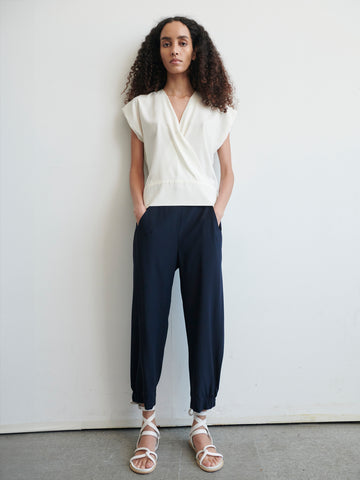 A person with long curly hair stands against a plain white background. They are wearing a white sleeveless wrap top, navy blue cropped Abi Pants by Zero + Maria Cornejo with cinched ankles, and white strappy sandals. Their hands are in the pockets of their pants, made from stretch silk charmeuse, and they are looking straight ahead.