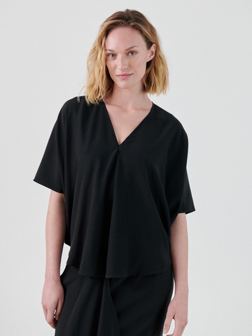 A person with shoulder-length blonde hair is wearing a loose-fitting black Circle Top with short sleeves, crafted from stretch silk charmeuse by Zero + Maria Cornejo. They are standing against a plain white background, slightly smiling with their right hand resting casually by their side.