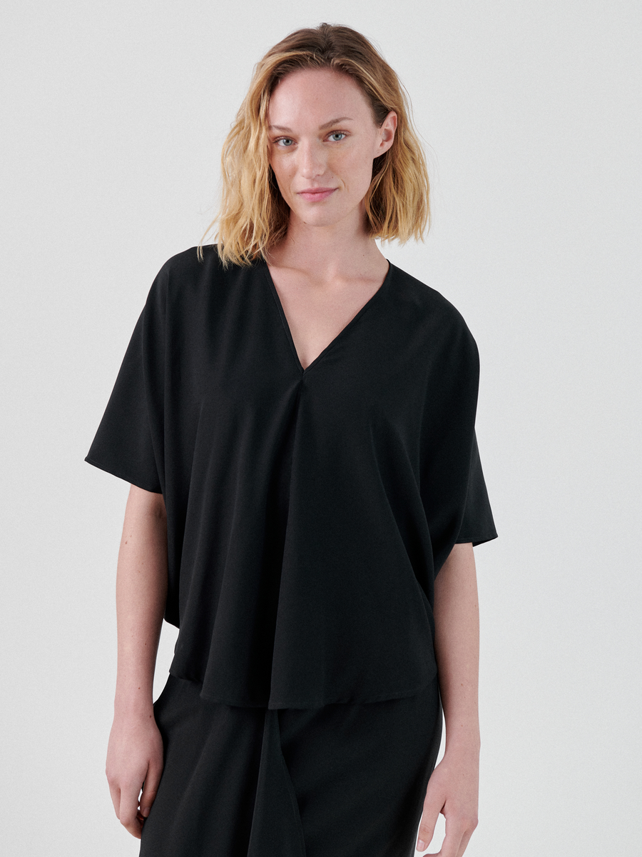 A person with shoulder-length blonde hair is wearing a loose-fitting black Circle Top with short sleeves, crafted from stretch silk charmeuse by Zero + Maria Cornejo. They are standing against a plain white background, slightly smiling with their right hand resting casually by their side.