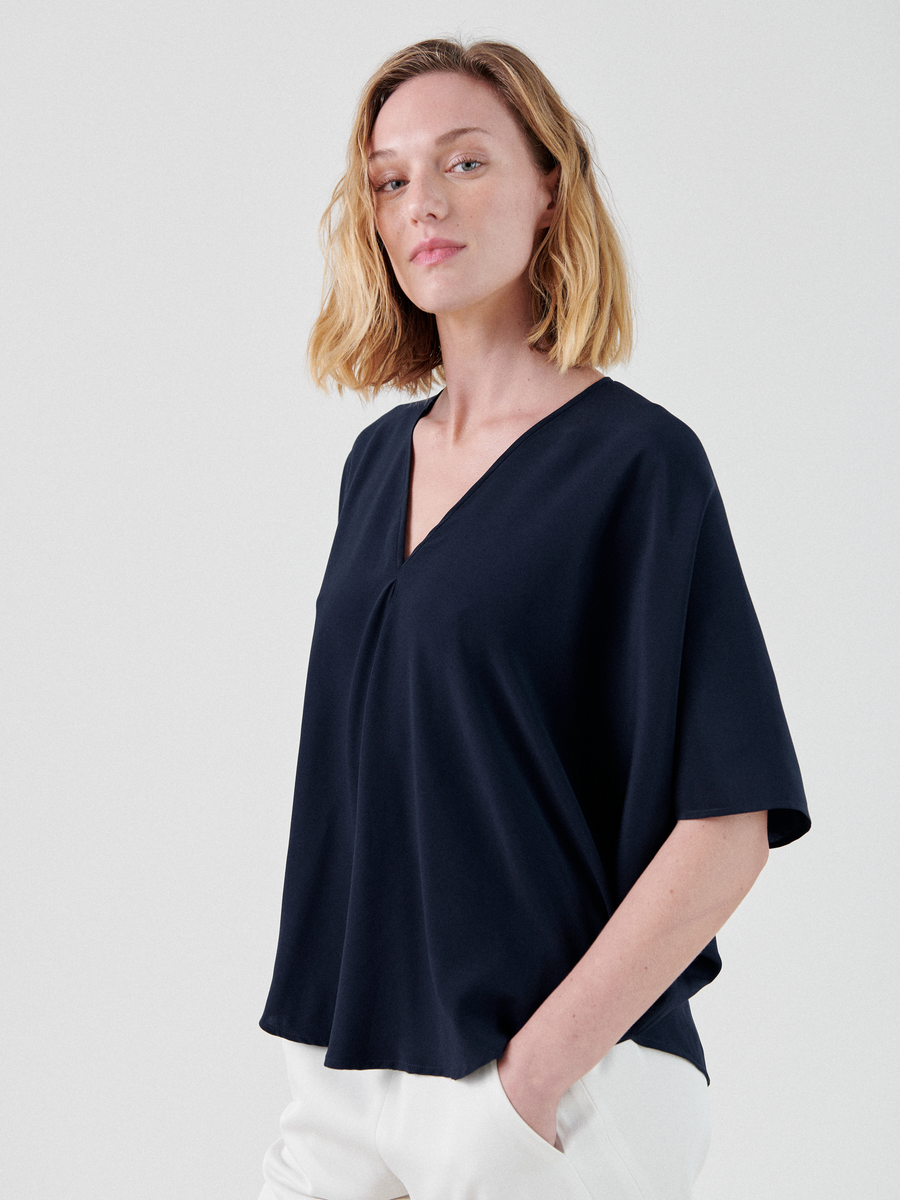 A person with shoulder-length blonde hair is standing against a plain background, wearing a loose-fitting, dark blue V-neck blouse and white pants. Made in New York by Zero + Maria Cornejo, this Circle Top adds an extra touch of elegance. They are gazing confidently at the camera with a relaxed posture.