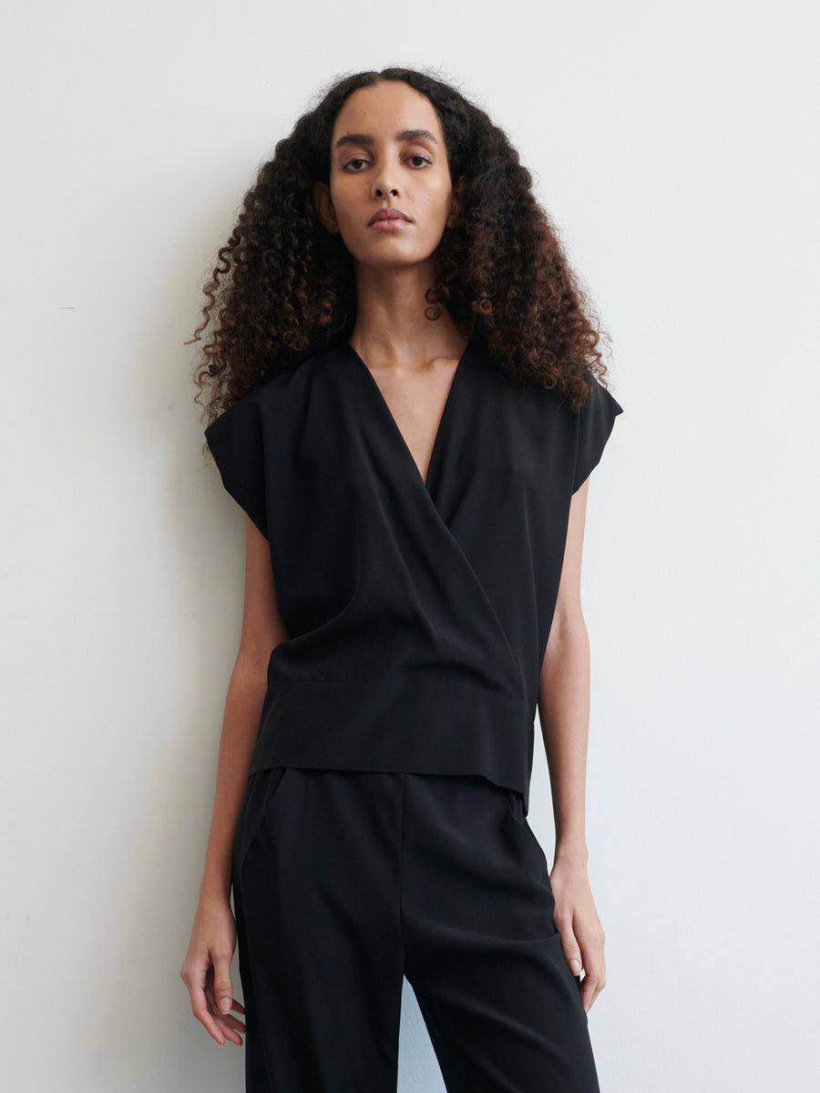 A person with long, curly hair wearing a Wrap Mido Top by Zero + Maria Cornejo and matching black pants stands against a plain white background. The person looks towards the camera with a neutral expression.