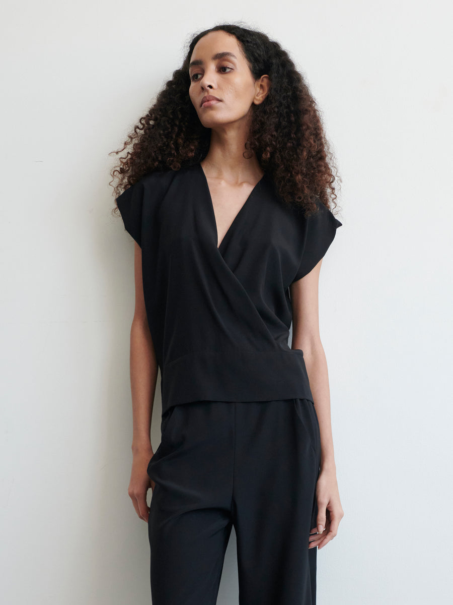 A person with long, curly hair wears a sleek black Wrap Mido Top by Zero + Maria Cornejo and matching black pants. They are standing against a plain white background and looking to the side with a neutral expression, their hands relaxed by their sides.