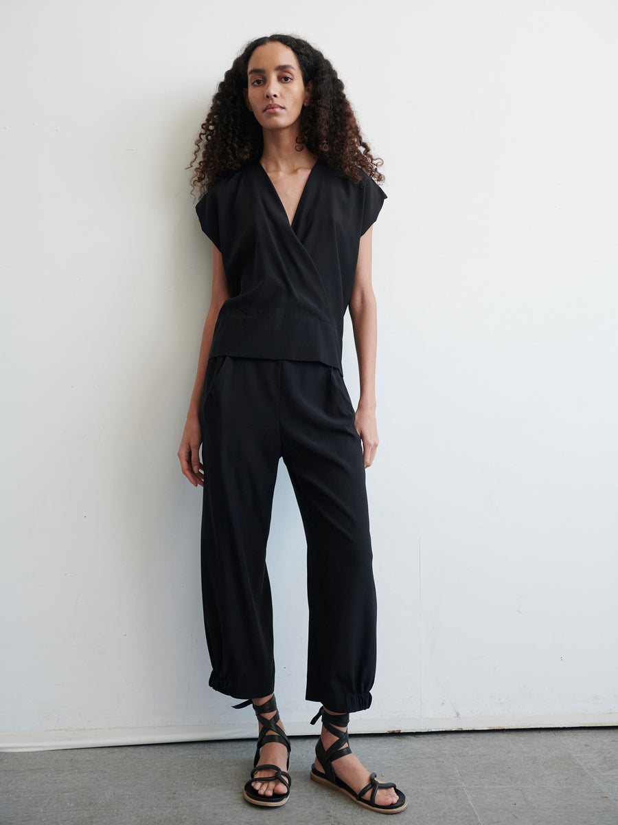 Person standing against a plain white wall, wearing a black Wrap Mido Top by Zero + Maria Cornejo and loose black pants cinched at the ankles. They have long, curly hair and are wearing black strappy sandals. The overall look is minimalist and modern.