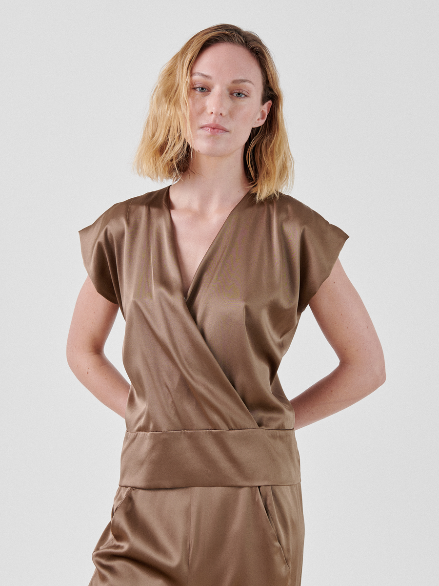 A person with shoulder-length blonde hair is wearing a brown Wrap Mido Top by Zero + Maria Cornejo made of natural fibers and matching trousers. The person stands against a plain light background, with their hands resting behind their back.