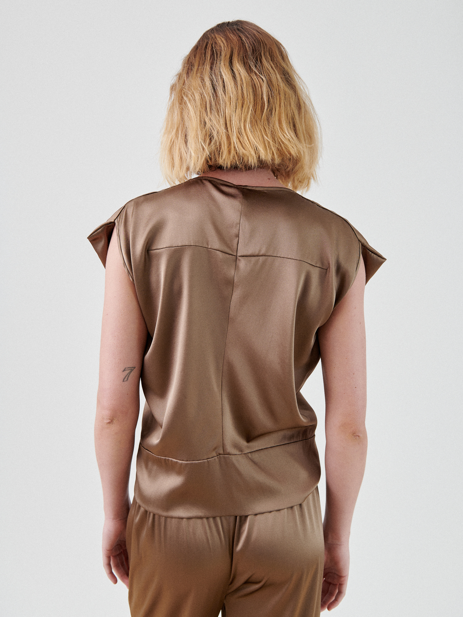 A person with shoulder-length, wavy blonde hair stands with their back to the camera, wearing a metallic bronze Wrap Mido Top by Zero + Maria Cornejo and matching pants. The background is plain and light-colored. A small tattoo is visible on the person's left forearm.