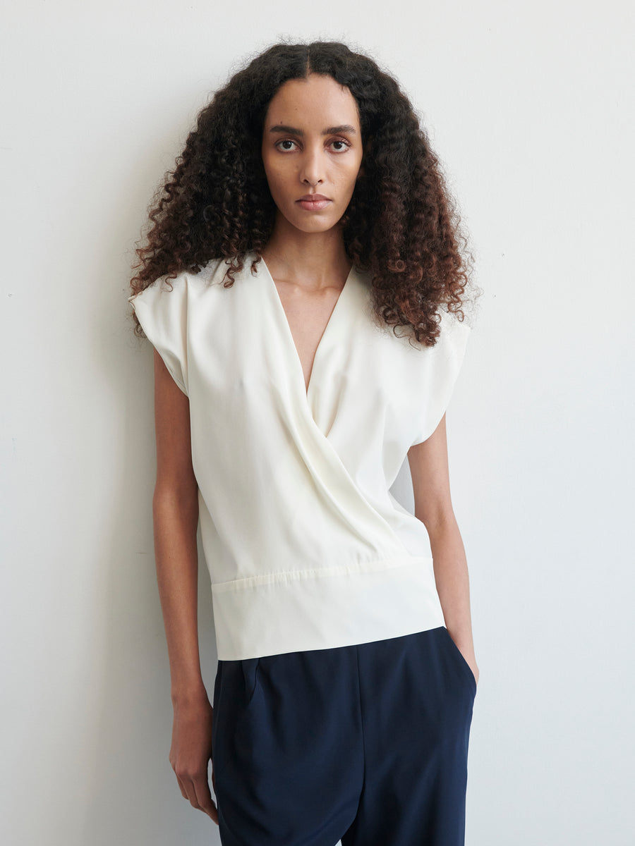 A person with long curly hair stands against a white background, looking directly at the camera with a neutral expression. They are wearing a sleeveless white silk charmeuse Wrap Mido Top by Zero + Maria Cornejo and dark navy blue pants. Their hands are in their pockets.