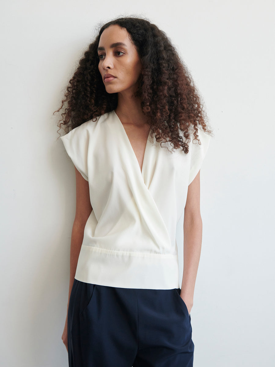 A woman with long curly hair wears a Zero + Maria Cornejo Wrap Mido Top and dark pants, standing against a plain light-colored wall with one hand in her pocket. She gazes thoughtfully to the side.