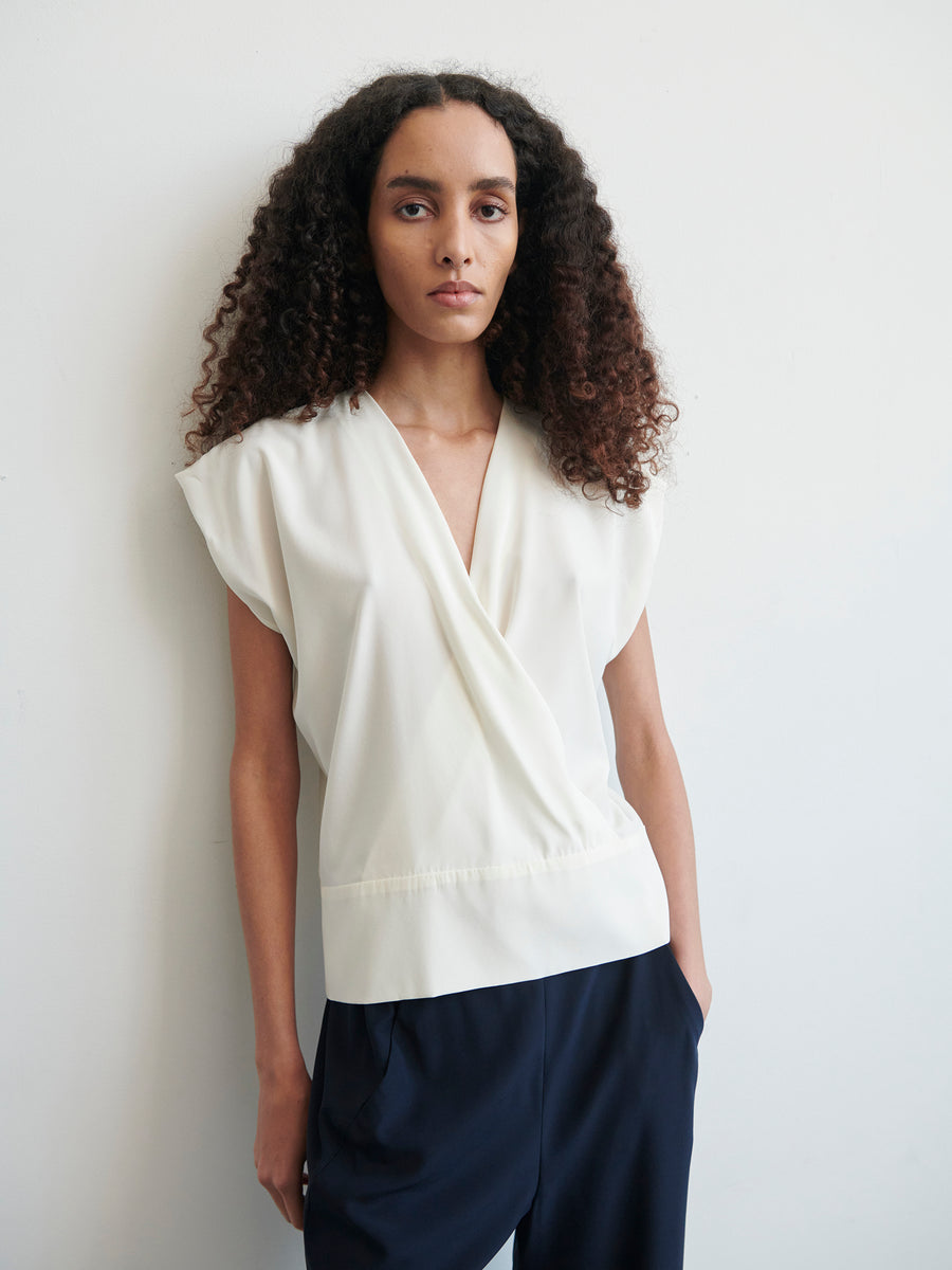 A woman with long, curly hair is standing against a plain, light-colored background. She is wearing a white Wrap Mido Top by Zero + Maria Cornejo with draped detailing and dark blue trousers. Her hands are in her pockets, and she has a neutral expression on her face.