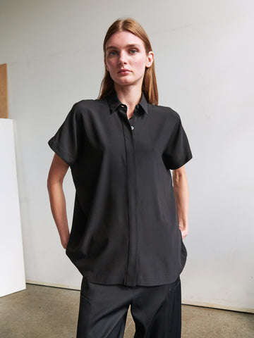 A person with long brown hair, wearing a loose-fitting black short-sleeve button down collared Gaban Shirt from Zero + Maria Cornejo and black pants, stands indoors against a plain white wall, looking slightly off-camera with hands in their pockets.