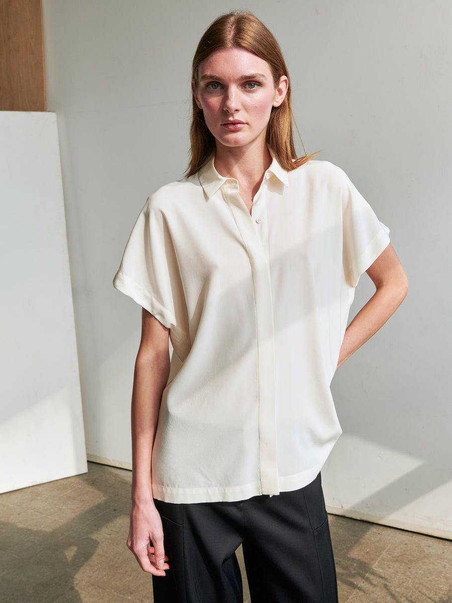 A woman stands indoors against a light-colored wall, wearing the Zero + Maria Cornejo Gaban Shirt, a white short-sleeved button-up with a hidden button placket, paired with black pants. She has straight, shoulder-length hair and one hand resting on her hip. Sunlight casts soft shadows in the background.