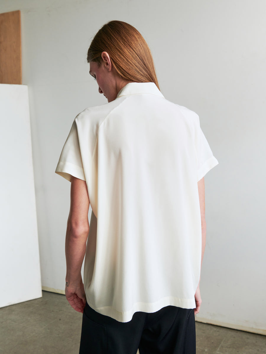 A person with long, light brown hair stands with their back to the camera, donning a Gaban Shirt by Zero + Maria Cornejo and black pants. The background features minimalistic, light-colored walls and a partial wooden board on the left.