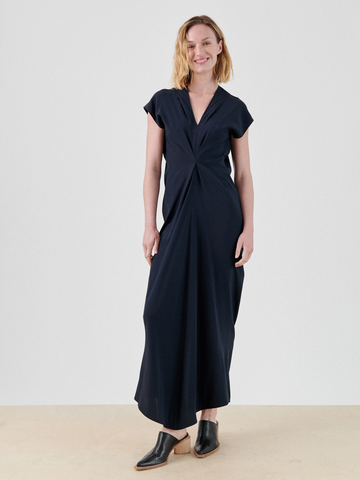 A woman with shoulder-length blonde hair is standing and smiling. She is wearing the Long Muse Dress by Zero + Maria Cornejo, a navy blue, short-sleeved, ankle-length draped silhouette dress with a central knot detail at the waist and black heeled mules. The background is plain and light-colored.