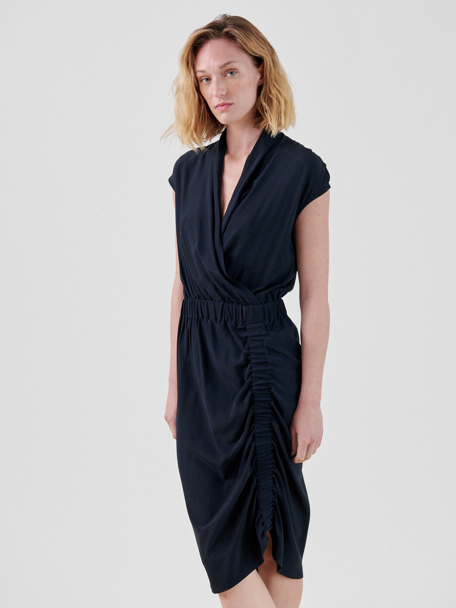 A person with shoulder-length blonde hair is wearing the Tasmeen Dress from Zero + Maria Cornejo, a dark blue, sleeveless knee-length dress featuring ruched detailing on the side. They stand against a plain, light gray background, looking directly at the camera with a neutral expression.