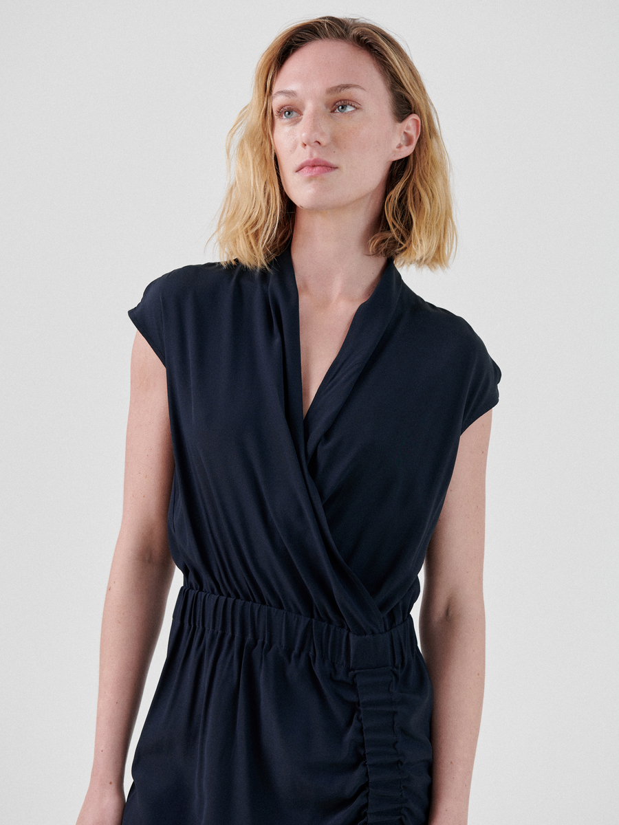 A person with shoulder-length blonde hair is wearing the Tasmeen Dress, a dark blue, sleeveless knee-length dress by Zero + Maria Cornejo, featuring a wrapped V-neck and gathered fabric at the waist. They are standing in front of a plain, light-colored background, looking slightly off to the side.