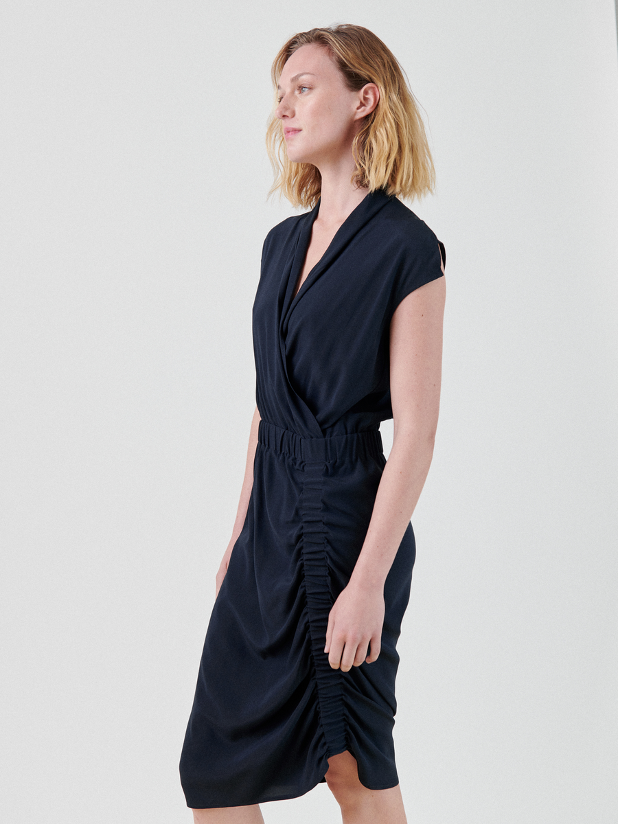 A woman with shoulder-length blonde hair is wearing the Tasmeen Dress by Zero + Maria Cornejo, a sleeveless, knee-length navy blue dress featuring a v-neck wrap-style neckline and ruched detailing along the skirt. She is standing and gazing slightly to the left against a plain white background.