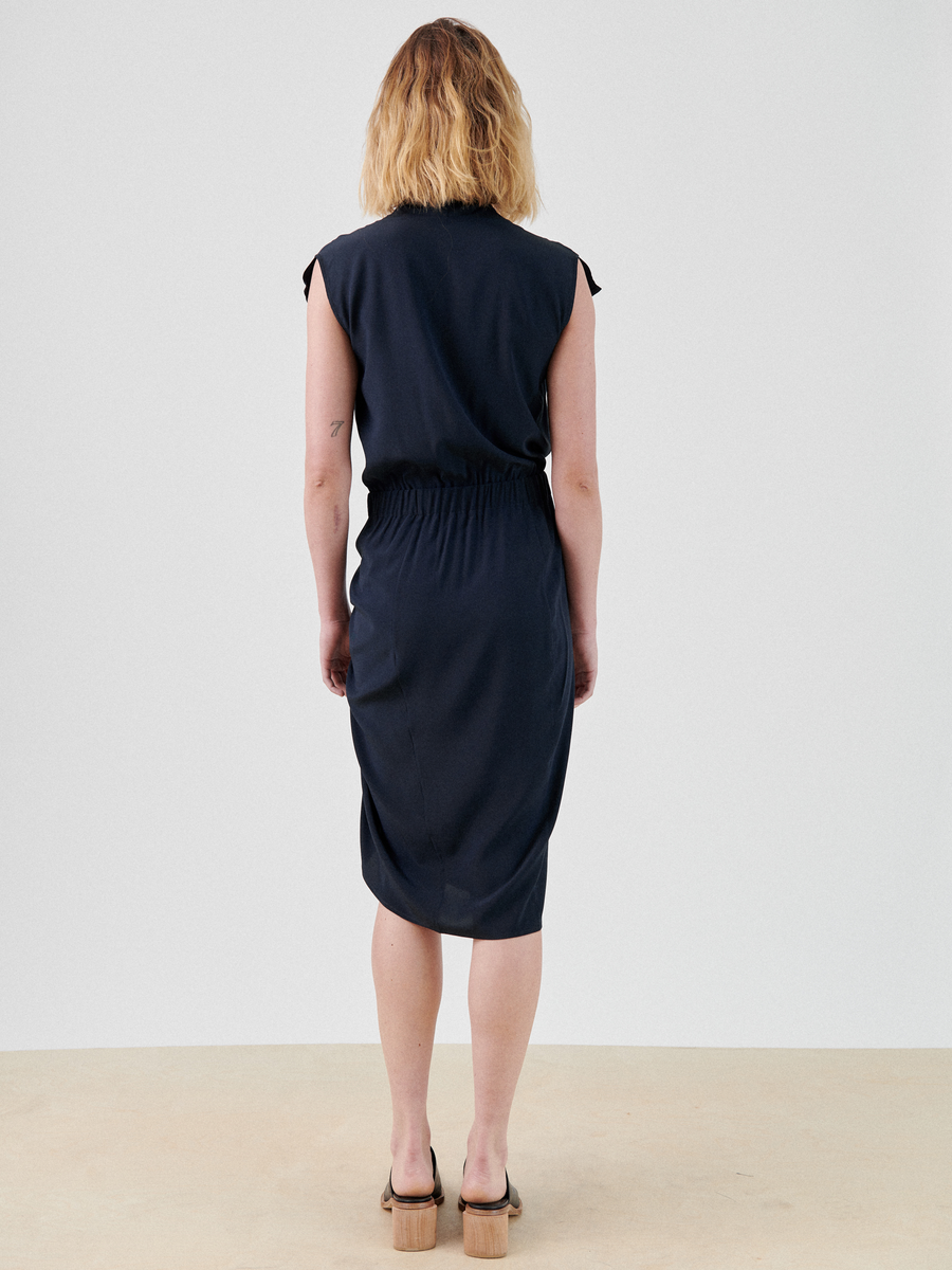 A person with shoulder-length blonde hair is standing with their back to the camera, wearing the knee-length Tasmeen Dress by Zero + Maria Cornejo, a black v-neck wrap dress with a cinched waist and black sandals. The background is a plain light gray.