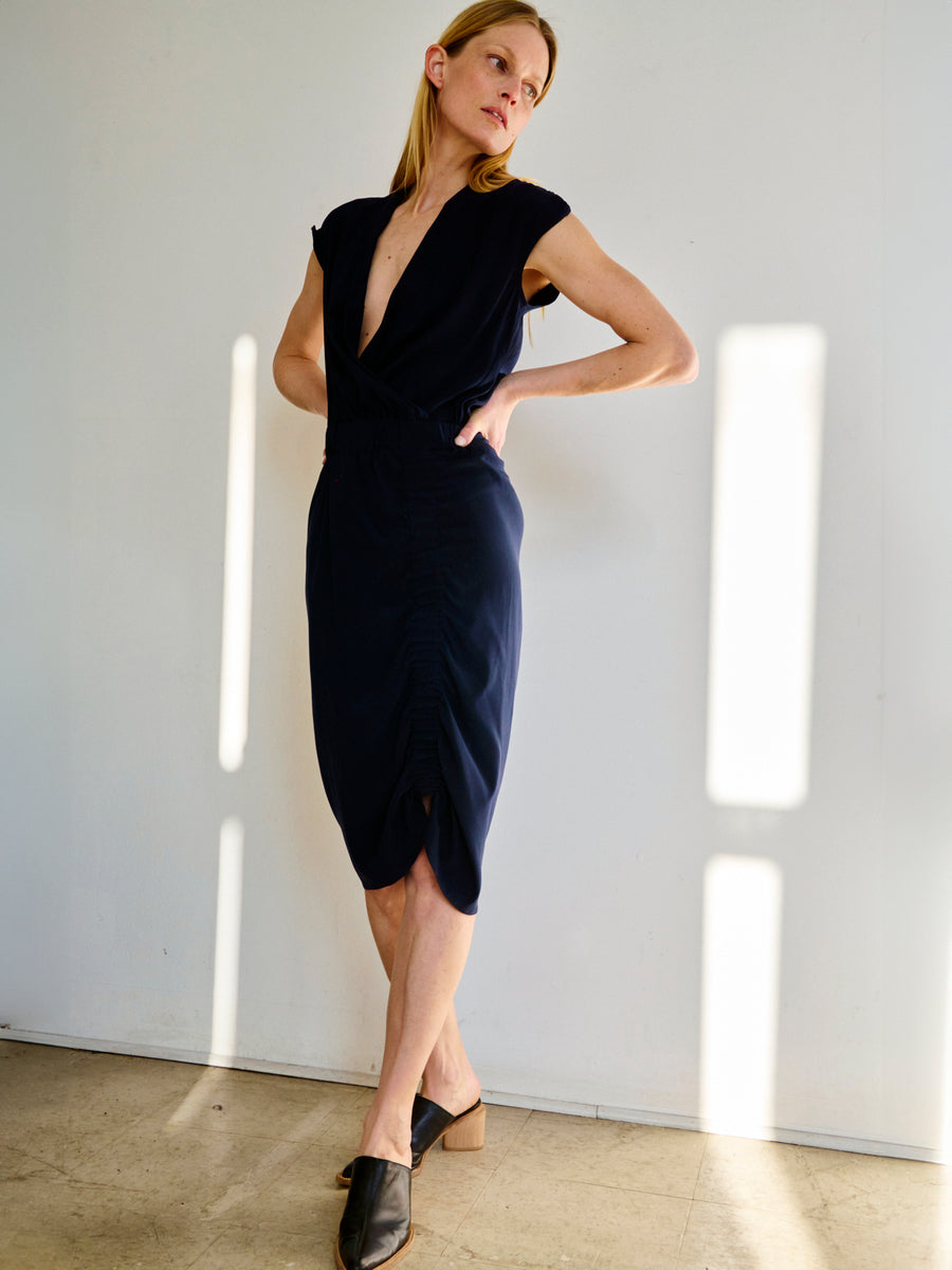 A woman stands against a plain white wall in a well-lit room, wearing the dark navy v-neck Tasmeen Dress by Zero + Maria Cornejo and black heeled mules. She has one hand on her hip and her blonde hair is tucked behind her ears. Sunlight casts shadows on the wall behind her.