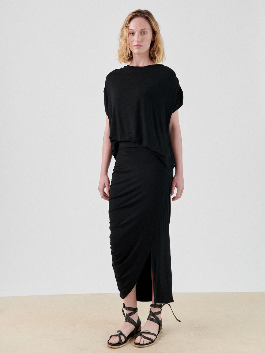 A person with shoulder-length blonde hair stands facing the camera, wearing a loose black top and an ankle-length Ruched Lui Skirt by Zero + Maria Cornejo with a side slit. They are also wearing black strappy sandals. The background is plain white, and the floor is beige.