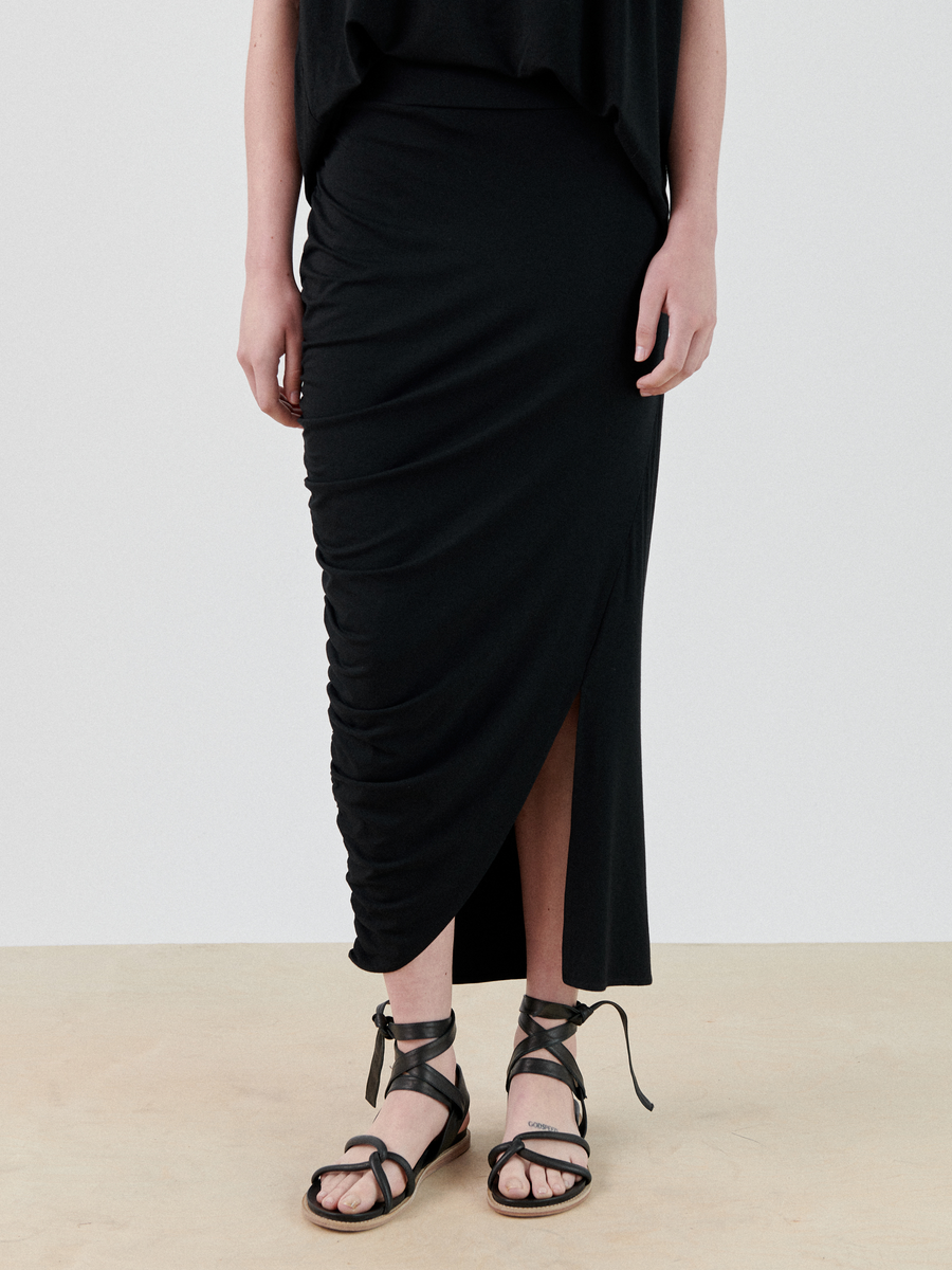 A person wearing a long black Ruched Lui Skirt from Zero + Maria Cornejo with an asymmetrical slit stands on a beige floor. They are also sporting black strappy sandals that wrap around the ankles. The upper part of the person is out of the frame.