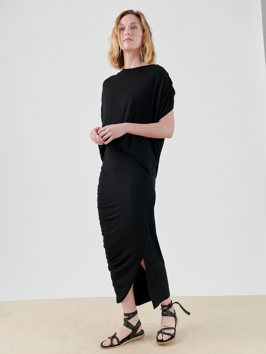 A person with shoulder-length blonde hair stands against a plain backdrop, wearing a loose-fitting black top and a Ruched Lui Skirt by Zero + Maria Cornejo with an asymmetrical silhouette. They have black strappy sandals on their feet and are looking slightly upward with a confident expression.