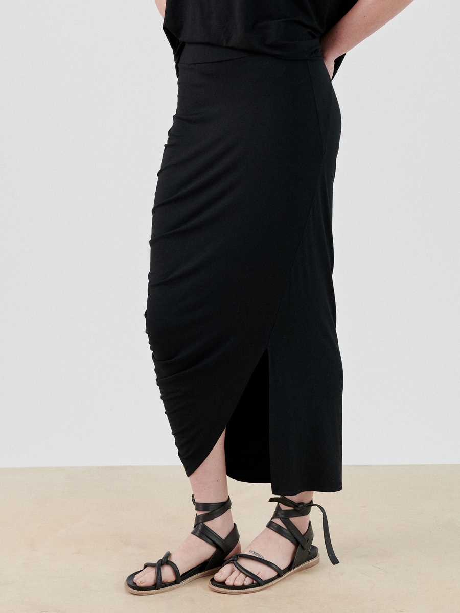 A person wearing a black, Ruched Lui Skirt by Zero + Maria Cornejo and strappy black sandals stands on a sandy surface. Only the lower half of their body and part of their hands resting on their hips are visible.