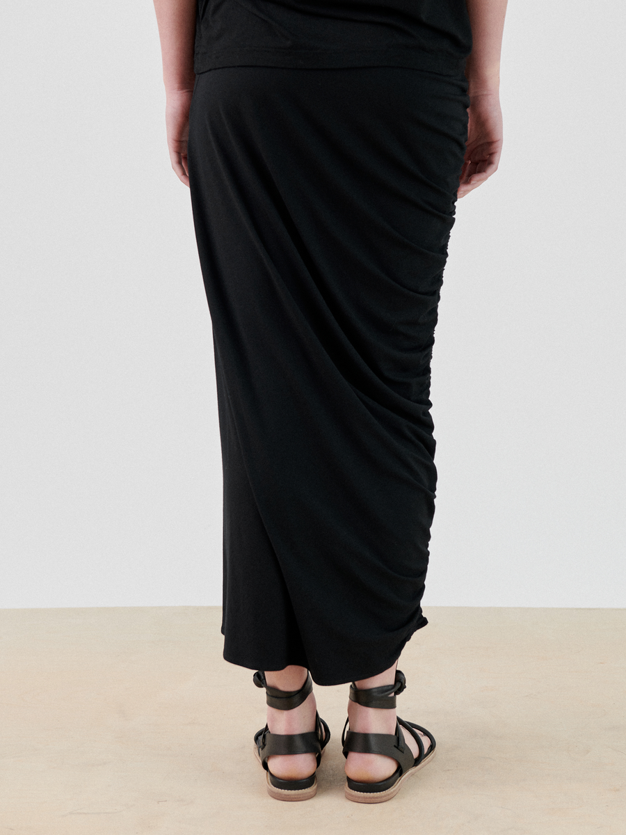 The image shows the lower back view of a person wearing a black Ruched Lui Skirt by Zero + Maria Cornejo that reaches the ankles and black sandals. The skirt has ruched detail on one side, creating an asymmetrical silhouette. The person is standing on a light-colored surface against a plain white background.