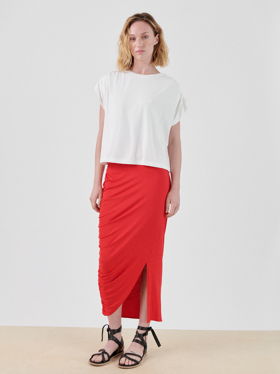 A person with shoulder-length blonde hair is wearing a white short-sleeved top and a red Ruched Lui Skirt by Zero + Maria Cornejo with an asymmetrical silhouette and front slit. They stand against a plain, light-colored background, wearing black sandals with ankle ties.