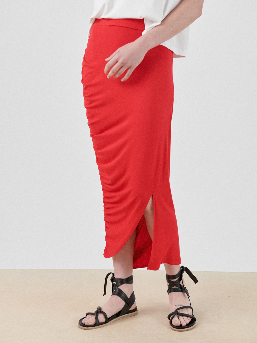 A person is wearing a red Ruched Lui Skirt from Zero + Maria Cornejo with a slit above the knee, crafted from sustainable fabric. They are also wearing a white top and black lace-up flat sandals. The background is plain.