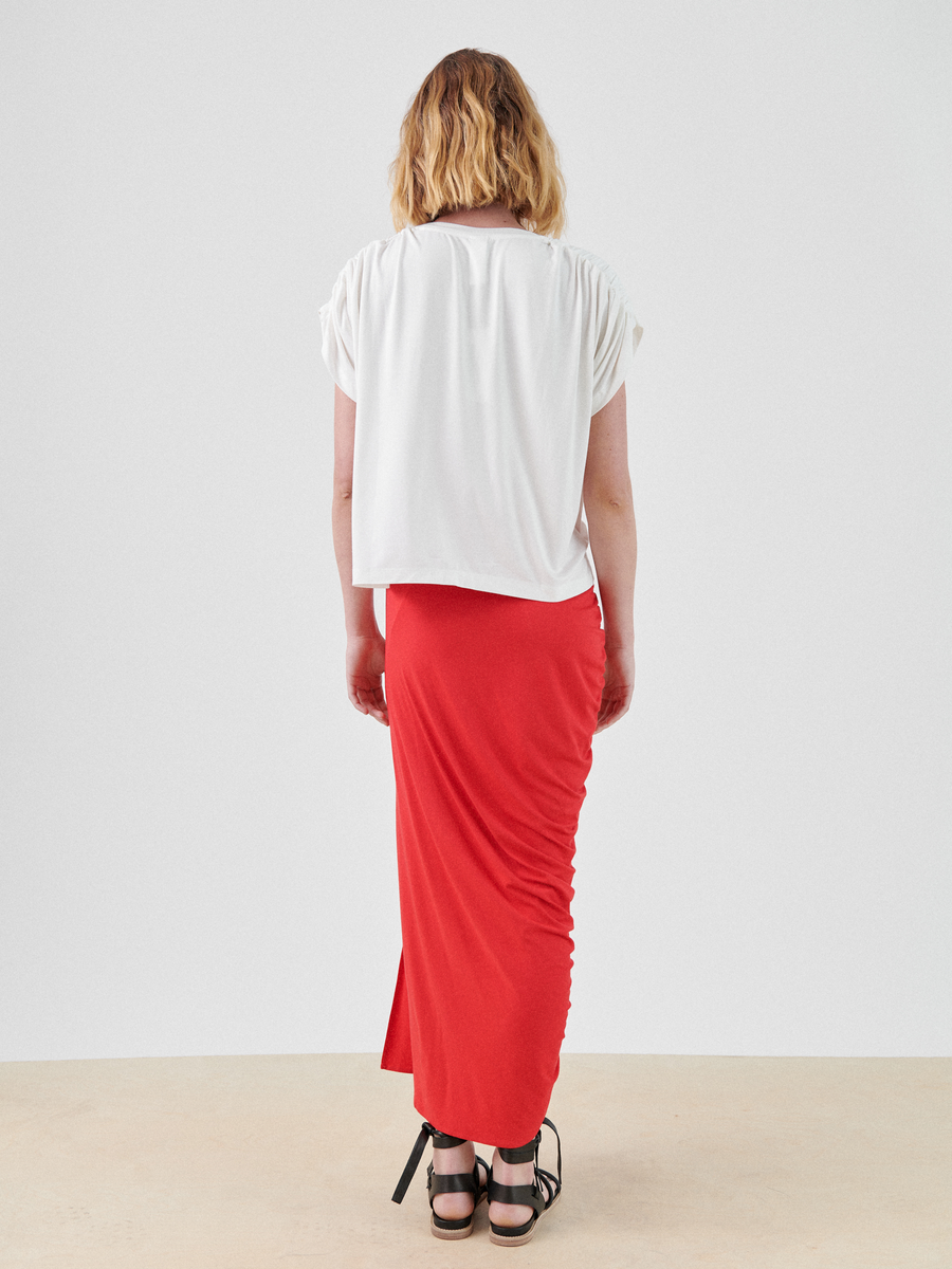 A person with shoulder-length blonde hair is standing facing away from the camera. They are wearing a white short-sleeve top and a long red Ruched Lui Skirt by Zero + Maria Cornejo paired with black strappy sandals. The background is plain white, and the floor is light-colored.