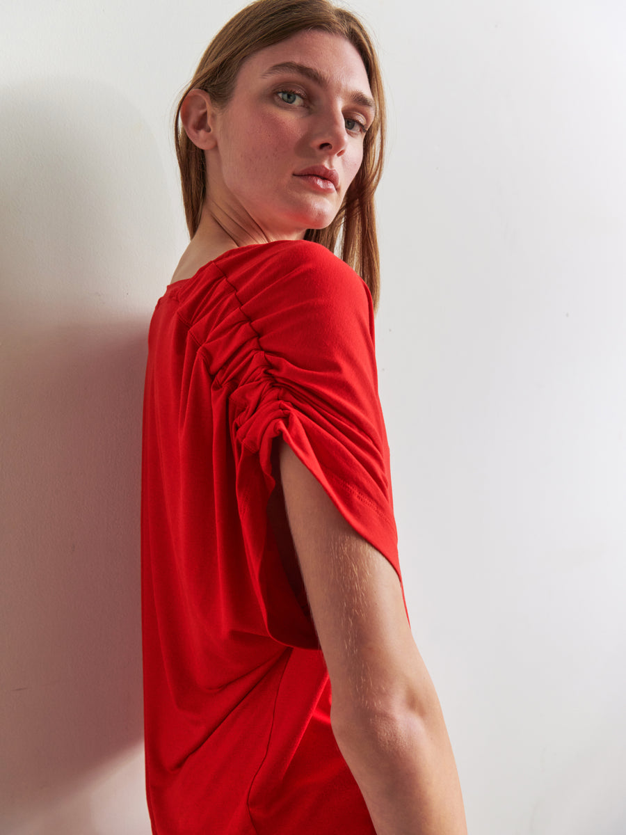 A person with long hair is standing against a white wall, turned slightly to the side, wearing the Ruched Muscle Tank by Zero + Maria Cornejo in a bright red shade. The top features ruched sleeves and is made from biodegradable fabric. The expression is neutral, and the lighting provides a soft, natural look to the scene.