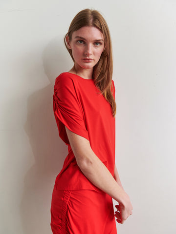 A person with long brown hair, wearing a red Ruched Muscle Tank from Zero + Maria Cornejo, made of biodegradable fabric stands against a plain white background. They have a neutral expression and are slightly turned to the side, with their arms resting at their sides. The tank features ruched sleeves and sides, complementing the round neck design.