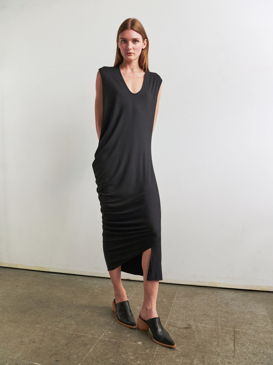 A person with long hair stands against a plain white wall, wearing the Curve Lui Dress by Zero + Maria Cornejo. The sleeveless, black, V-neck dress features an ankle-length design with an asymmetrical draped silhouette and a slight side slit. They have their hands behind their back and are paired with black open-back shoes that have wooden heels. The floor appears to be concrete.