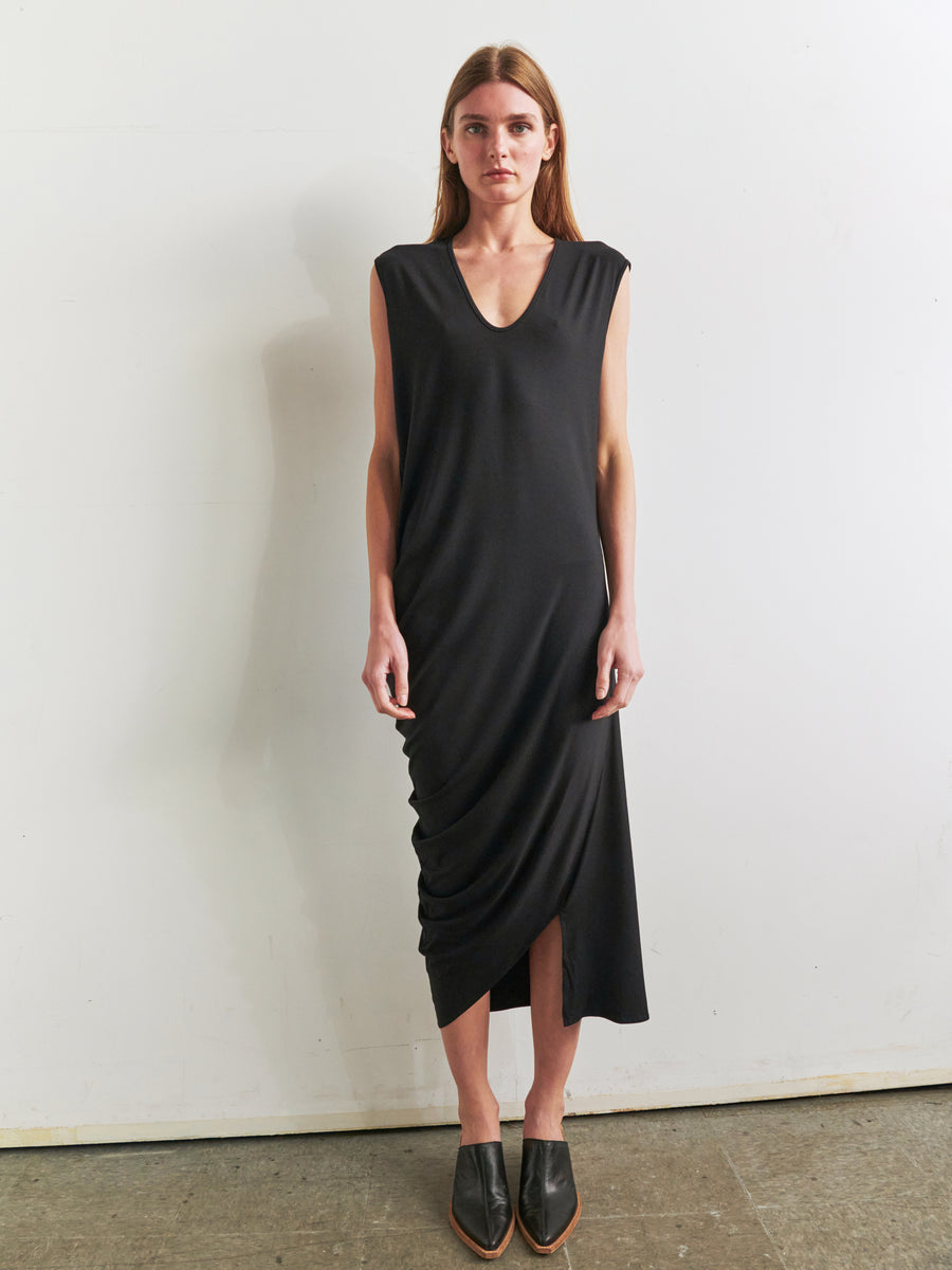 A woman stands against a plain white wall, wearing the Curve Lui Dress by Zero + Maria Cornejo. The sleeveless dress features an asymmetrical draped silhouette and a slightly ruched design on one side. The V-neck, black dress falls below her knees. She has long, straight hair and is wearing black, pointed-toe shoes.