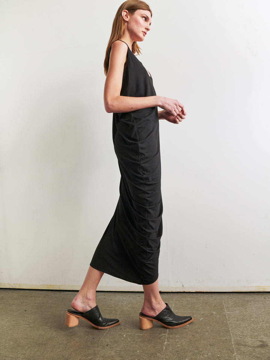 A person with long hair, dressed in the Curve Lui Dress by Zero + Maria Cornejo, walks sideways with their hands slightly lifted. The sleeveless black dress showcases an asymmetrical draped silhouette and is paired with black heeled mules. They are positioned against a plain white wall, and the floor appears to be a polished concrete surface.