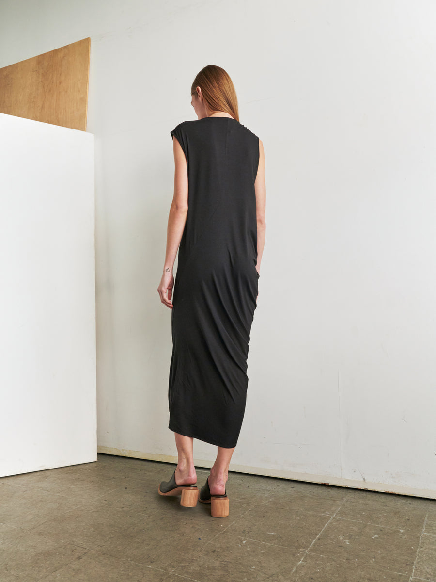 A person with long red hair is standing in a minimalistic room with their back to the camera. They are wearing the Curve Lui Dress by Zero + Maria Cornejo, a sleeveless, asymmetrical draped silhouette black dress made from biodegradable fabric, paired with brown platform sandals. A wooden panel leans against the white wall on the left side of the image.