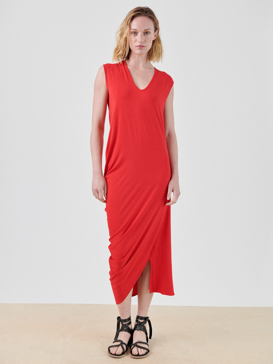 A person with shoulder-length blonde hair stands against a plain white background wearing the Curve Lui Dress by Zero + Maria Cornejo. The dress features a sleeveless red design with a V-neckline and an asymmetrical draped silhouette, crafted from biodegradable viscose. They are also wearing black strappy sandals.