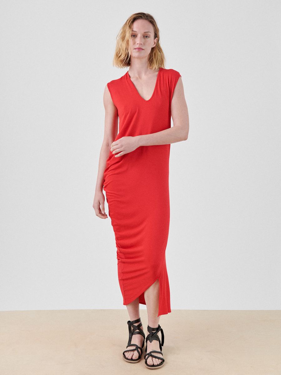 A person with shoulder-length blonde hair is wearing the Curve Lui Dress, a sleeveless red V-neck midi dress from Zero + Maria Cornejo, made from biodegradable viscose. They are standing against a plain white background and holding the side of the dress. They are wearing black strappy sandals with a low heel.