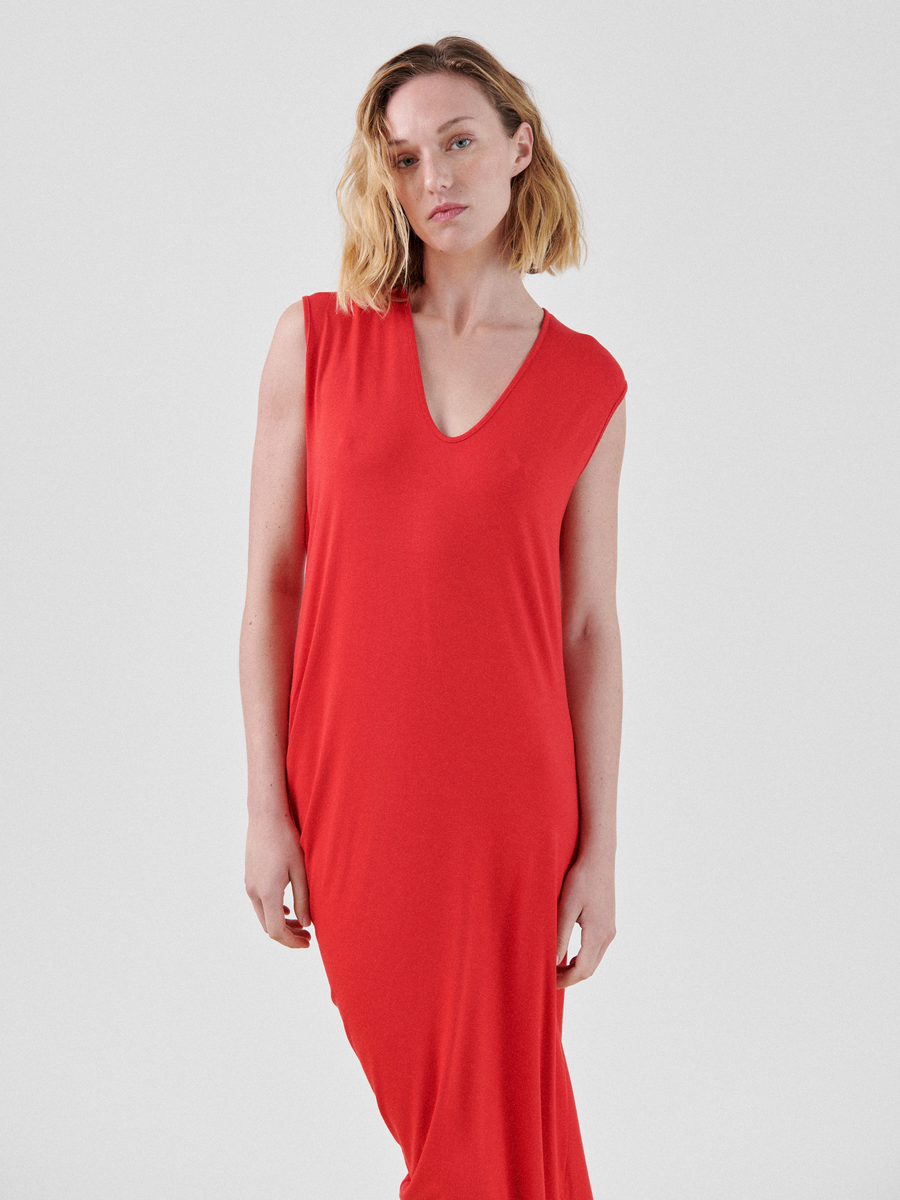 A woman with shoulder-length blonde hair stands against a plain background, wearing the Curve Lui Dress by Zero + Maria Cornejo. The sleeveless red dress is made from biodegradable viscose and features a V-neckline. She looks directly at the camera with a neutral expression, with one arm resting by her side.