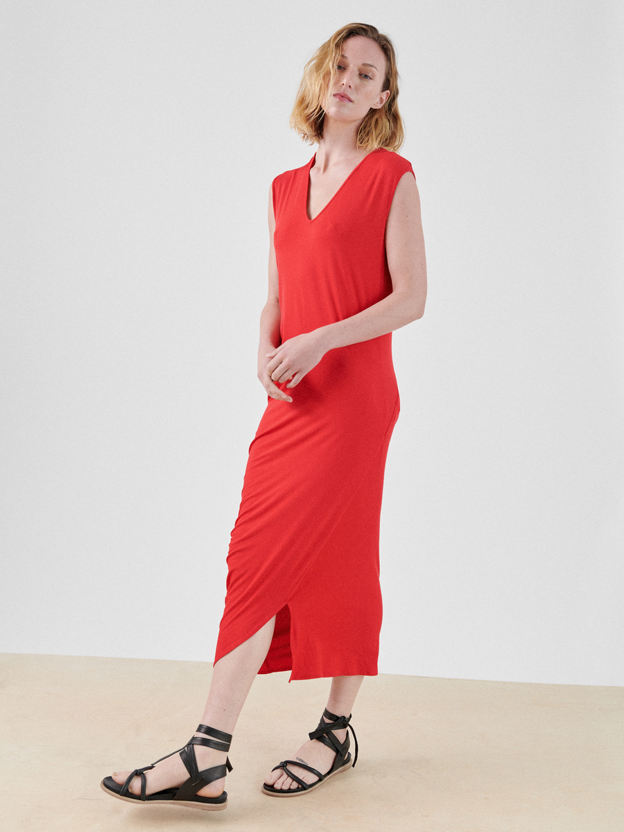 A person with shoulder-length hair wearing the Curve Lui Dress by Zero + Maria Cornejo stands against a plain white background. The sleeveless, bright red V-neck midi dress features an asymmetrical draped silhouette that adds flair to the biodegradable viscose fabric. They have black strappy sandals on and are slightly leaning to one side with one hand resting on the opposite wrist.