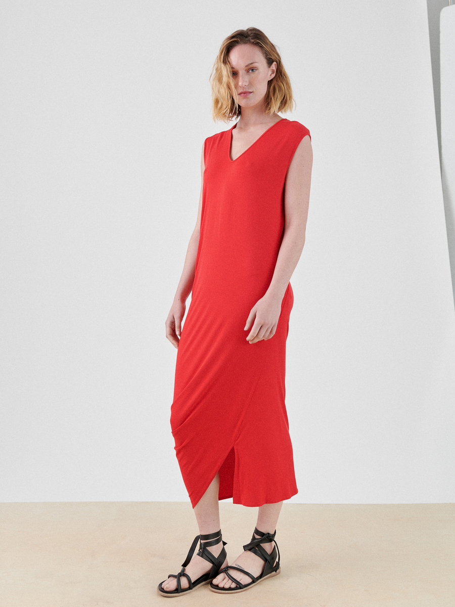 A woman with shoulder-length hair stands against a plain white background, showcasing the Curve Lui Dress by Zero + Maria Cornejo. The sleeveless, red midi dress features an asymmetrical draped silhouette and V-neck. She pairs it with black strappy sandals and looks directly at the camera with a neutral expression.