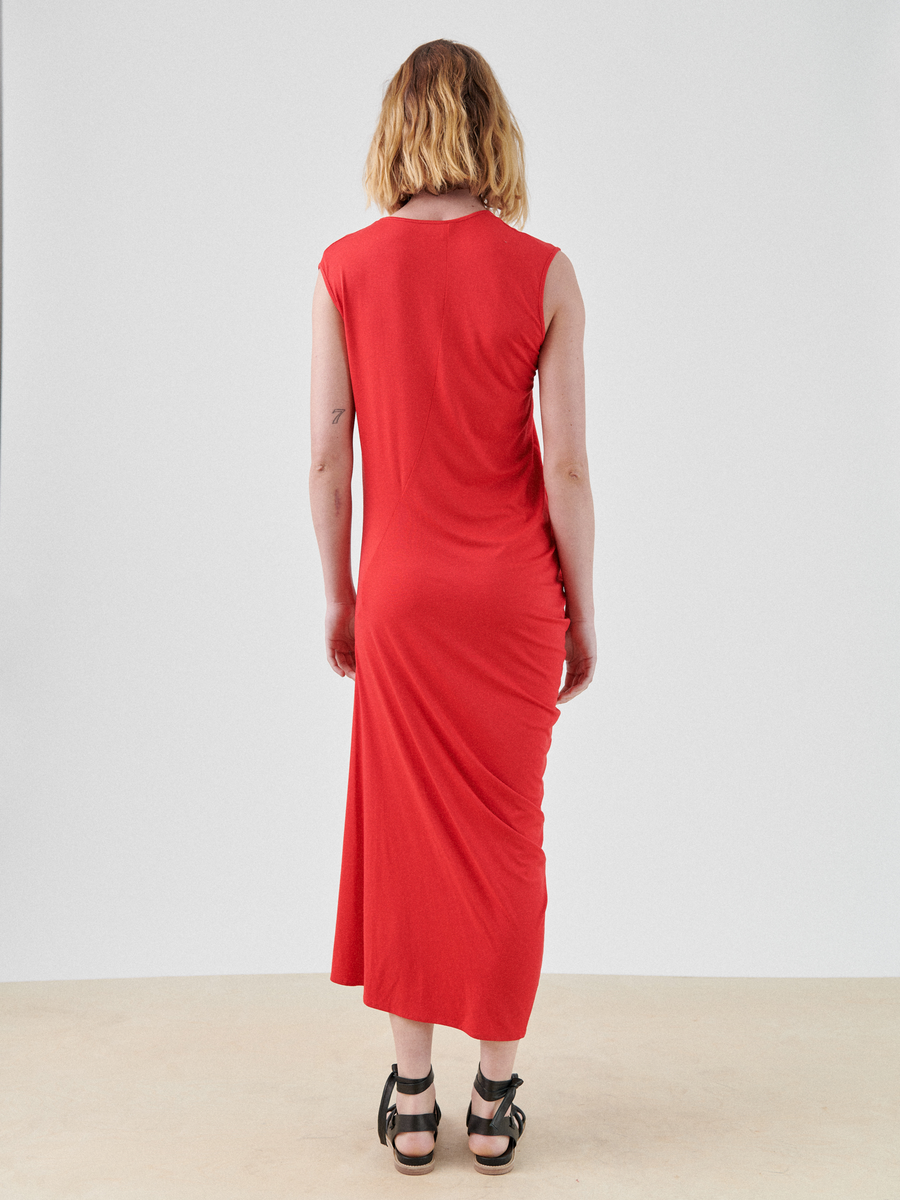 A person with blonde hair stands with their back to the camera, wearing the Curve Lui Dress by Zero + Maria Cornejo, showcasing an asymmetrical draped silhouette in a sleeveless red design, paired with black sandals. The background is plain and grayish-white.