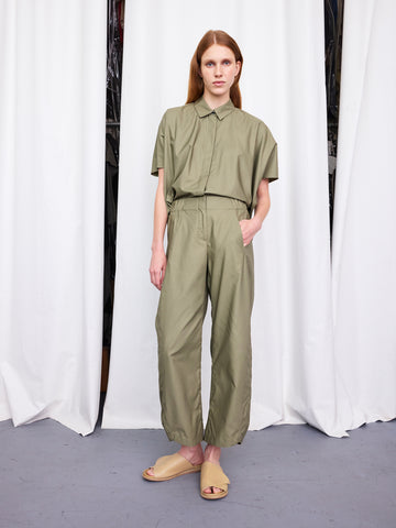 A person stands before white curtains wearing an olive green cotton broadcloth short-sleeve button-up shirt, the Zero + Maria Cornejo Aissa Cargo Pant in matching color, and beige open-toe slide sandals with hands in pockets.