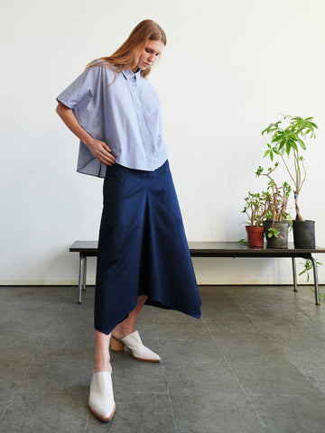 A woman with long red hair wears a loose light blue shirt, white shoes, and the Zero + Maria Cornejo Aissa Joi Skirt. She stands indoors near a bench with potted plants against a plain wall.